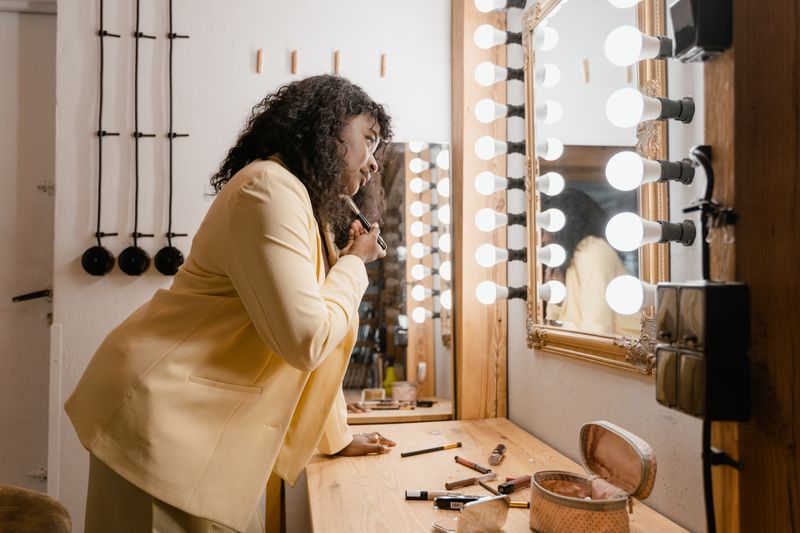 African American woman applying makeup at a vanity mirror with lights