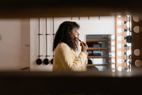 Free A woman applying makeup in front of a well-lit dressing room mirror, enhancing her look. Stock Photo