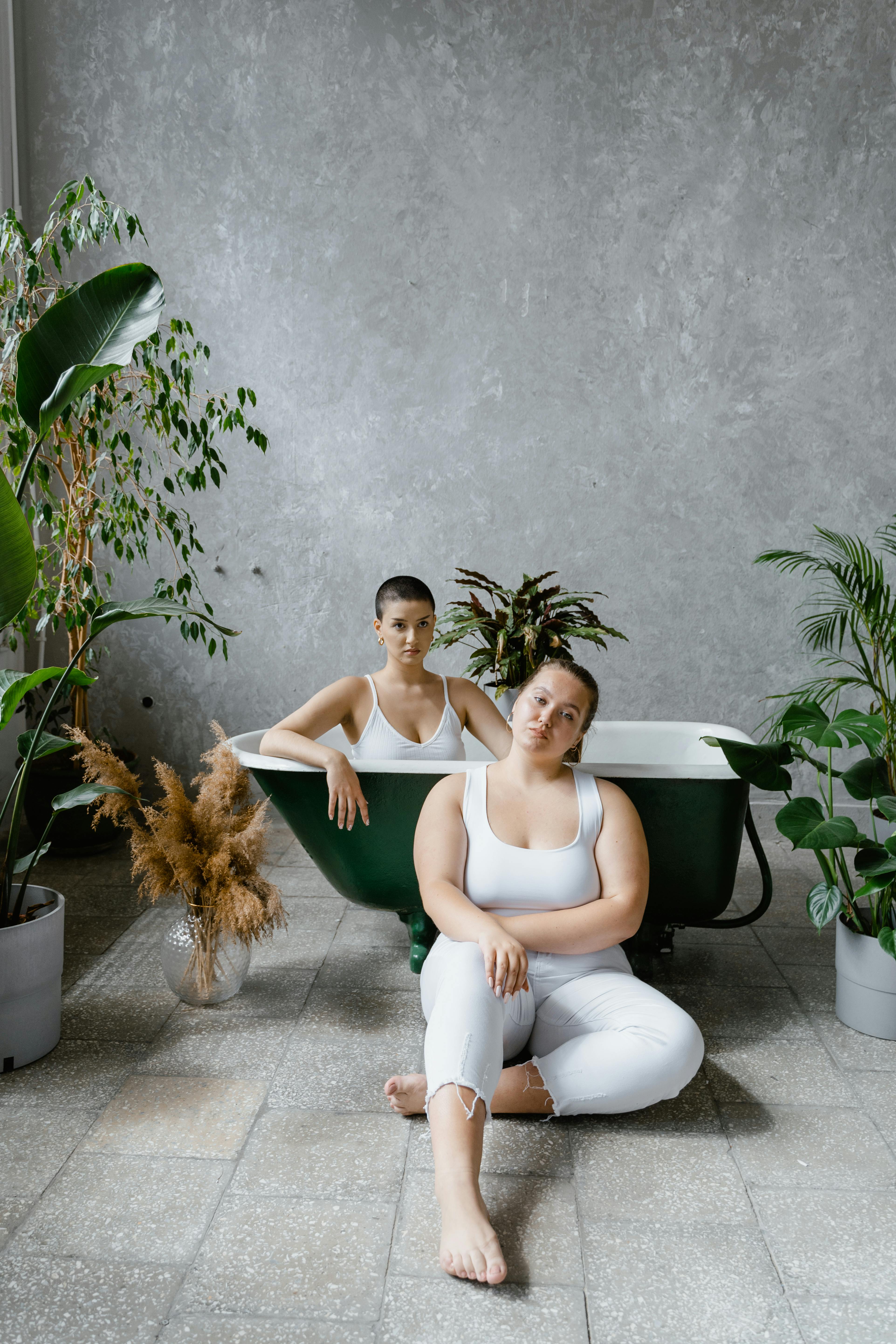 Two women in a bathtub surrounded by indoor plants, showcasing natural beauty and confidence.