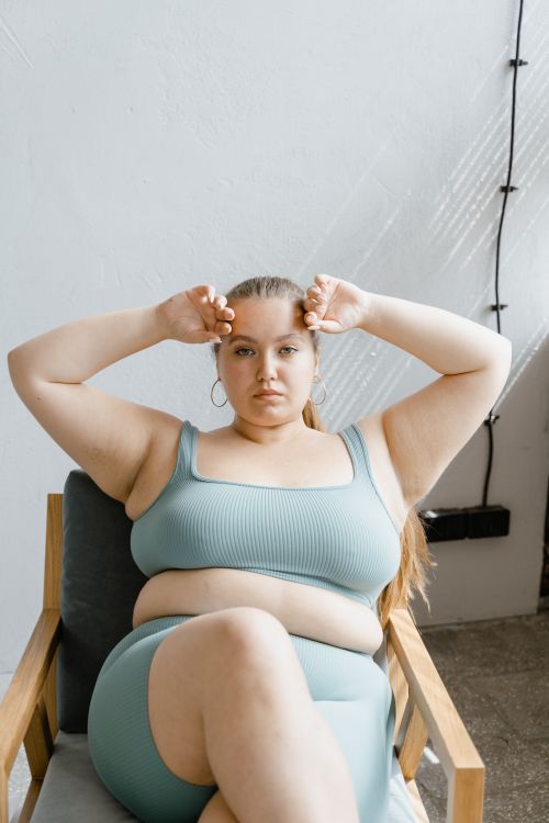 Free Portrait of a confident woman sitting in a chair indoors, wearing a teal bralette, with hands on head. Stock Photo