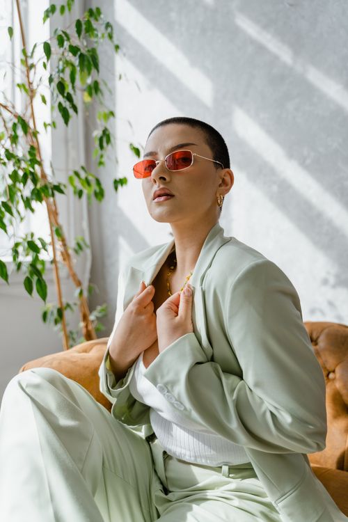Free Fashionable woman in a mint green suit with red sunglasses sitting indoors, exuding confidence. Stock Photo