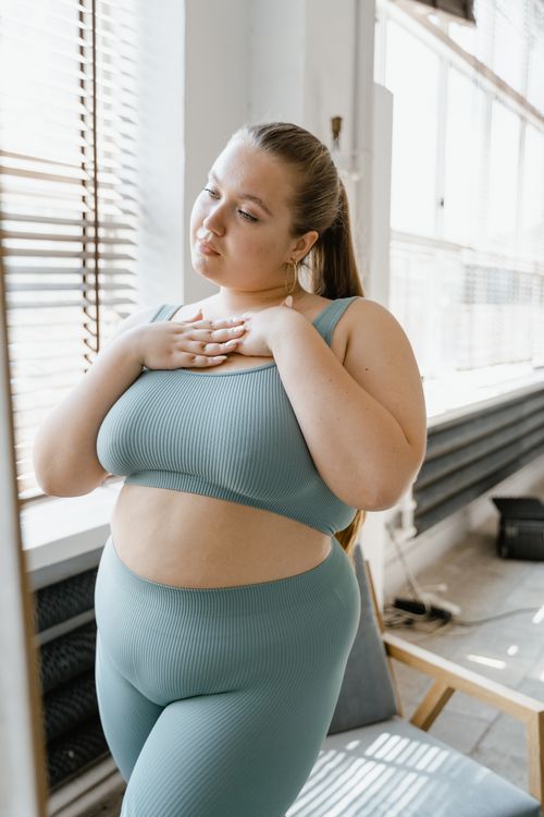 Free Plus-size woman in teal leisurewear standing by a window, exuding confidence and self-esteem. Stock Photo