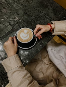 From above of crop anonymous female in trendy coat sitting at table with cup of coffee in cafe