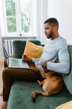 Young man works intently on laptop with dog by his side, sitting on a cozy sofa.