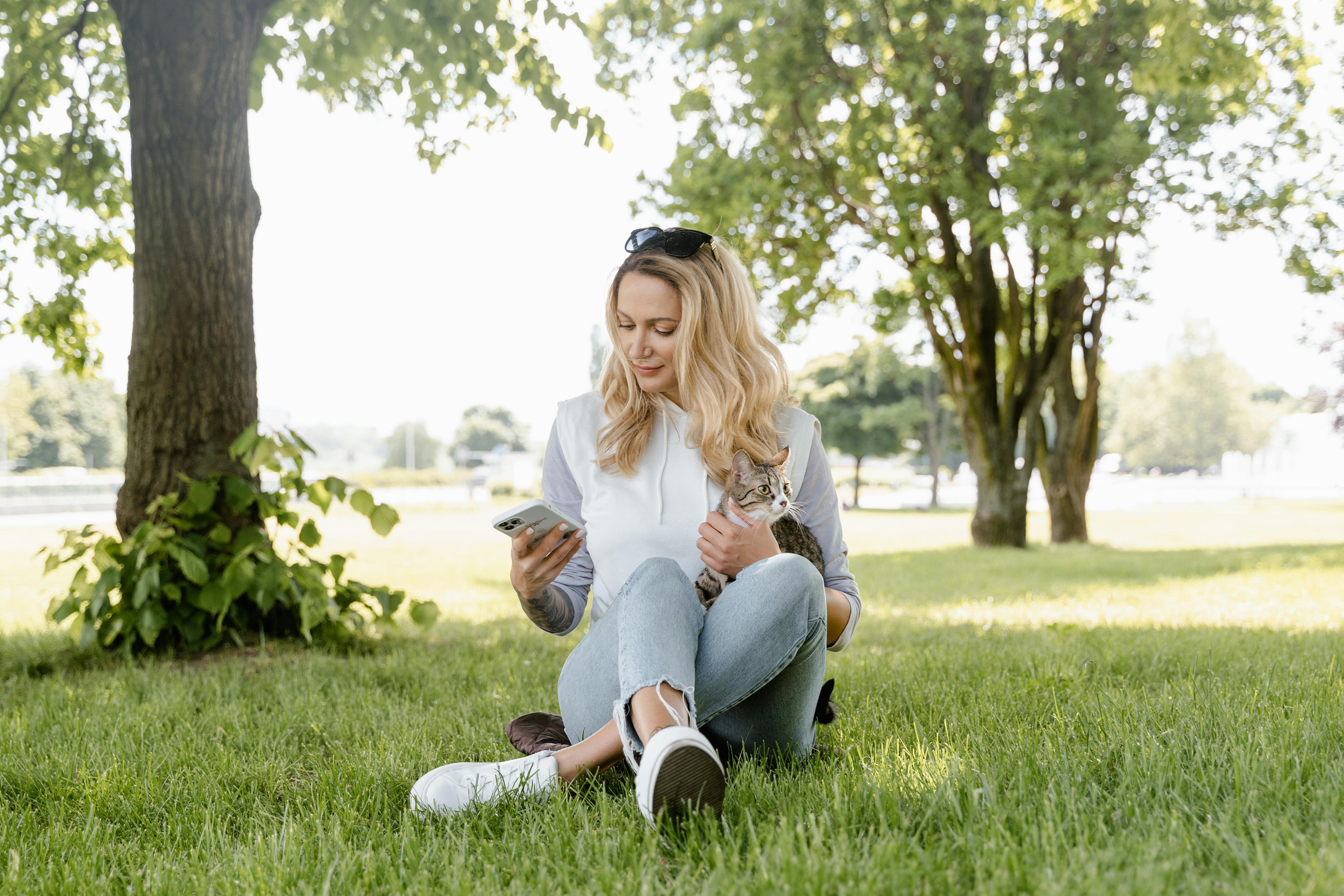 A woman sitting on grass, holding a cat and using her smartphone on a sunny day.