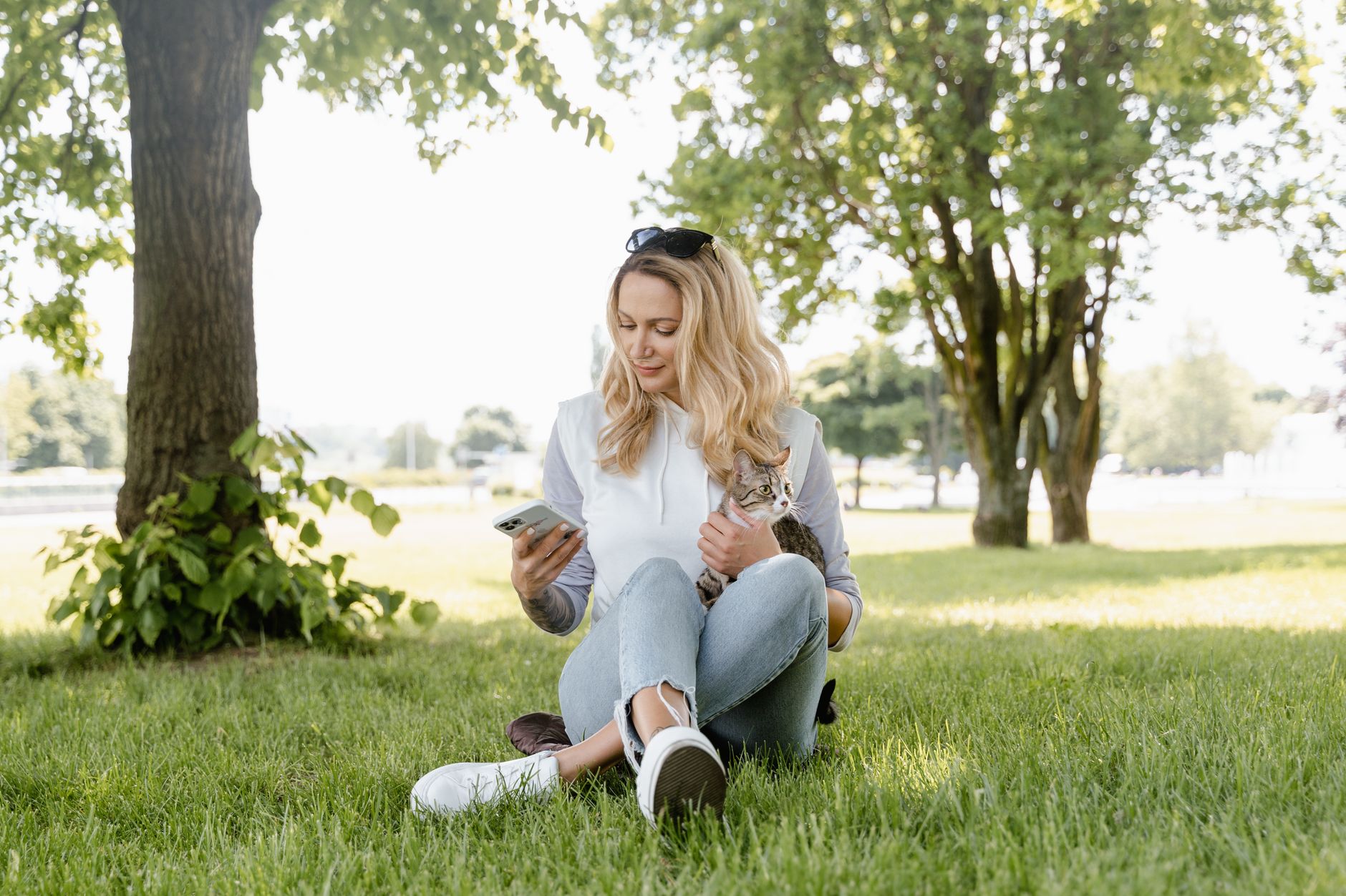 A woman sitting on grass, holding a cat and using her smartphone on a sunny day.