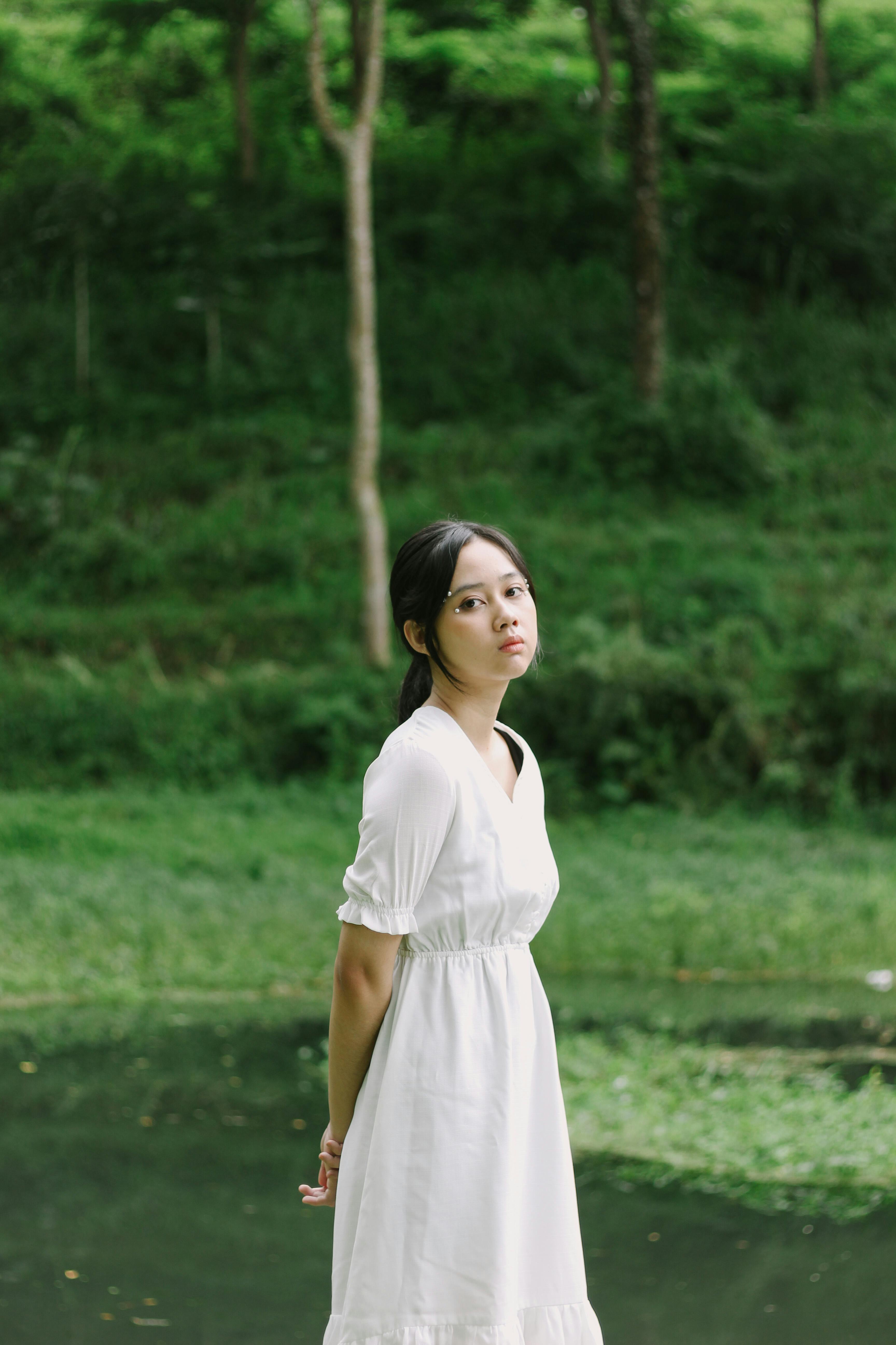 A serene portrait of a woman in a white dress amidst lush green nature, looking contemplative.