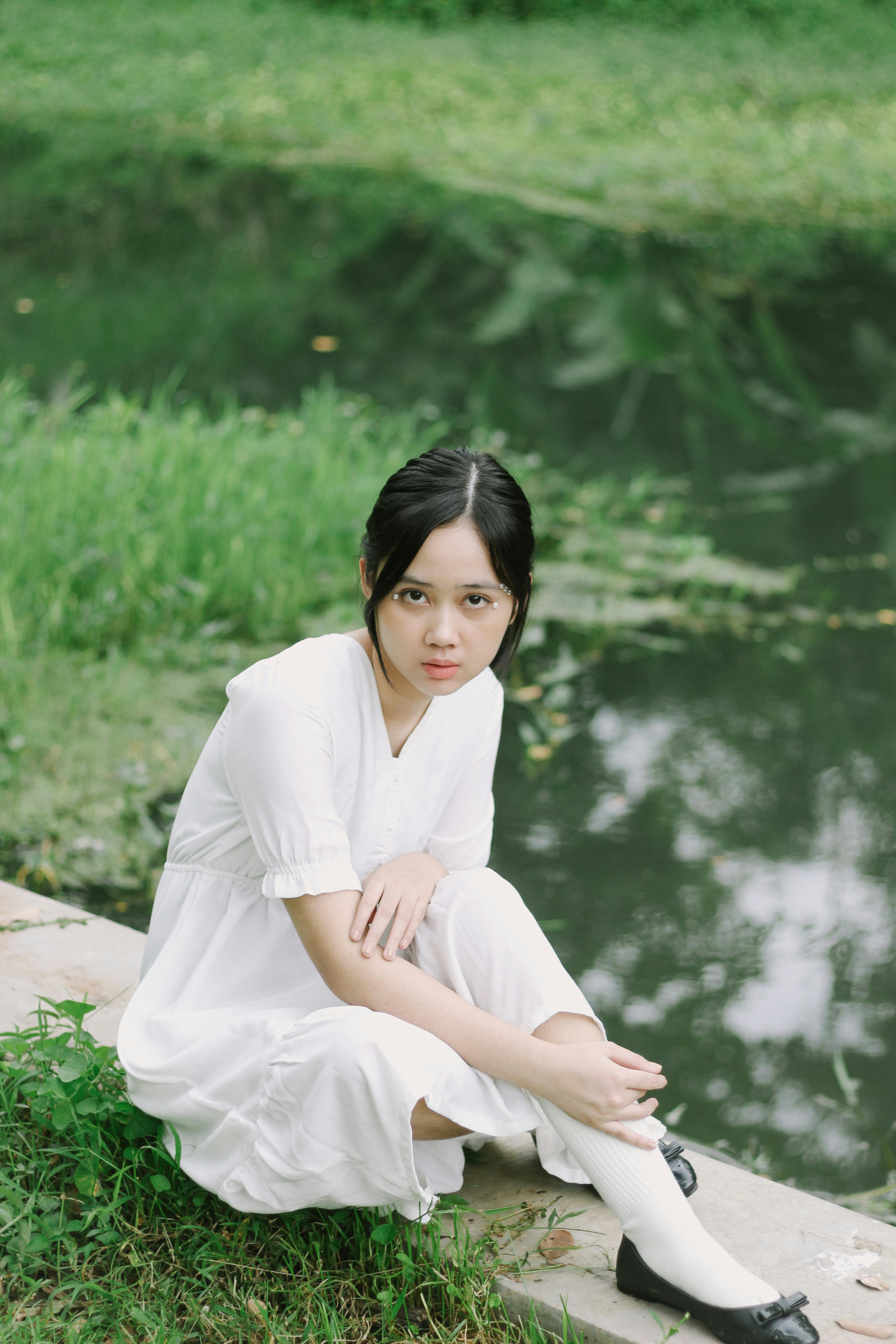 Asian woman in a white dress sitting by a tranquil river, exuding elegance.