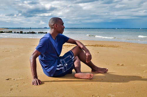 Free Man in Blue Shirt Sitting on Brown Sand near the Beach Stock Photo