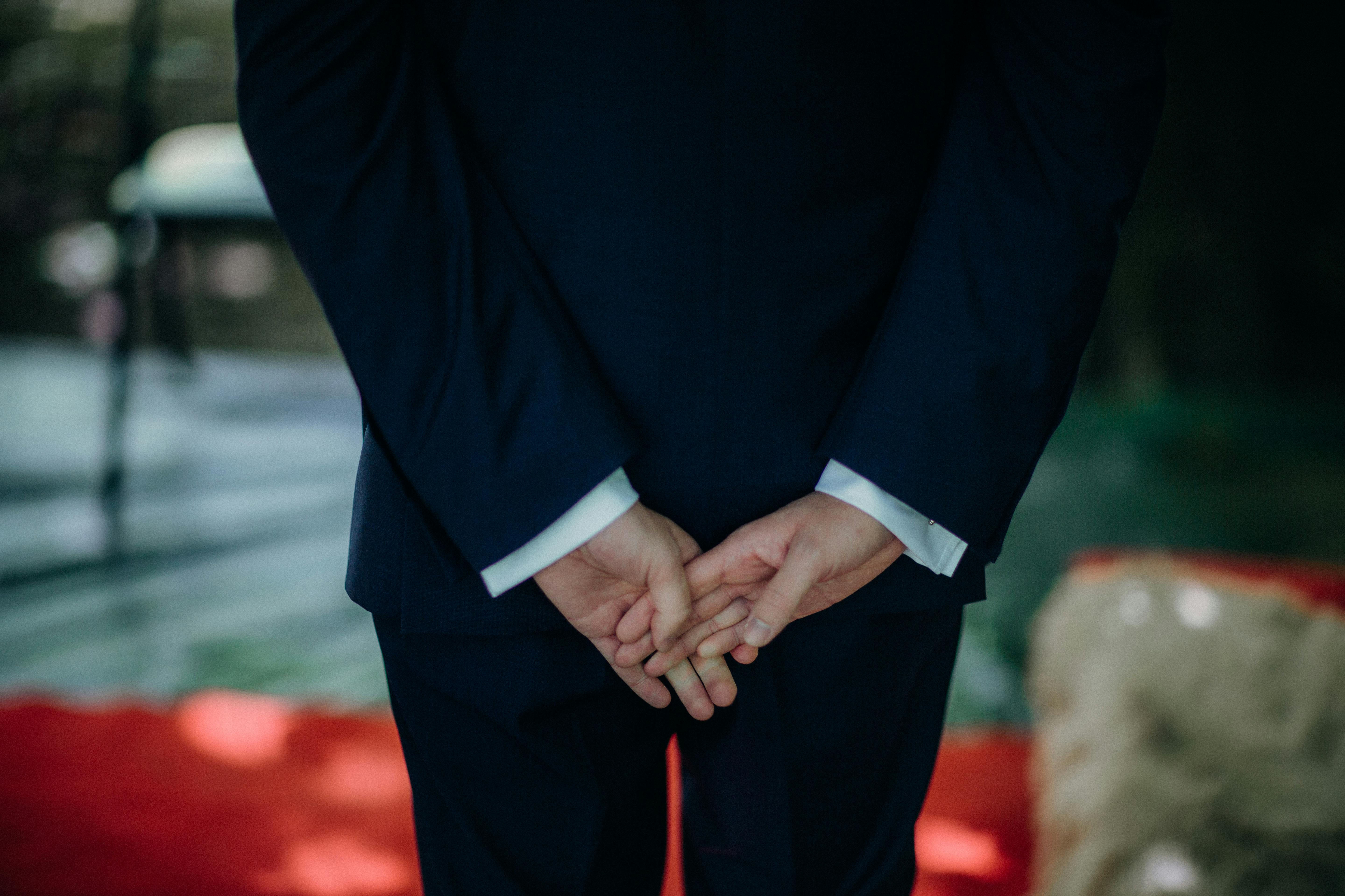 Close-up of a man in a suit standing with hands behind his back indoors.