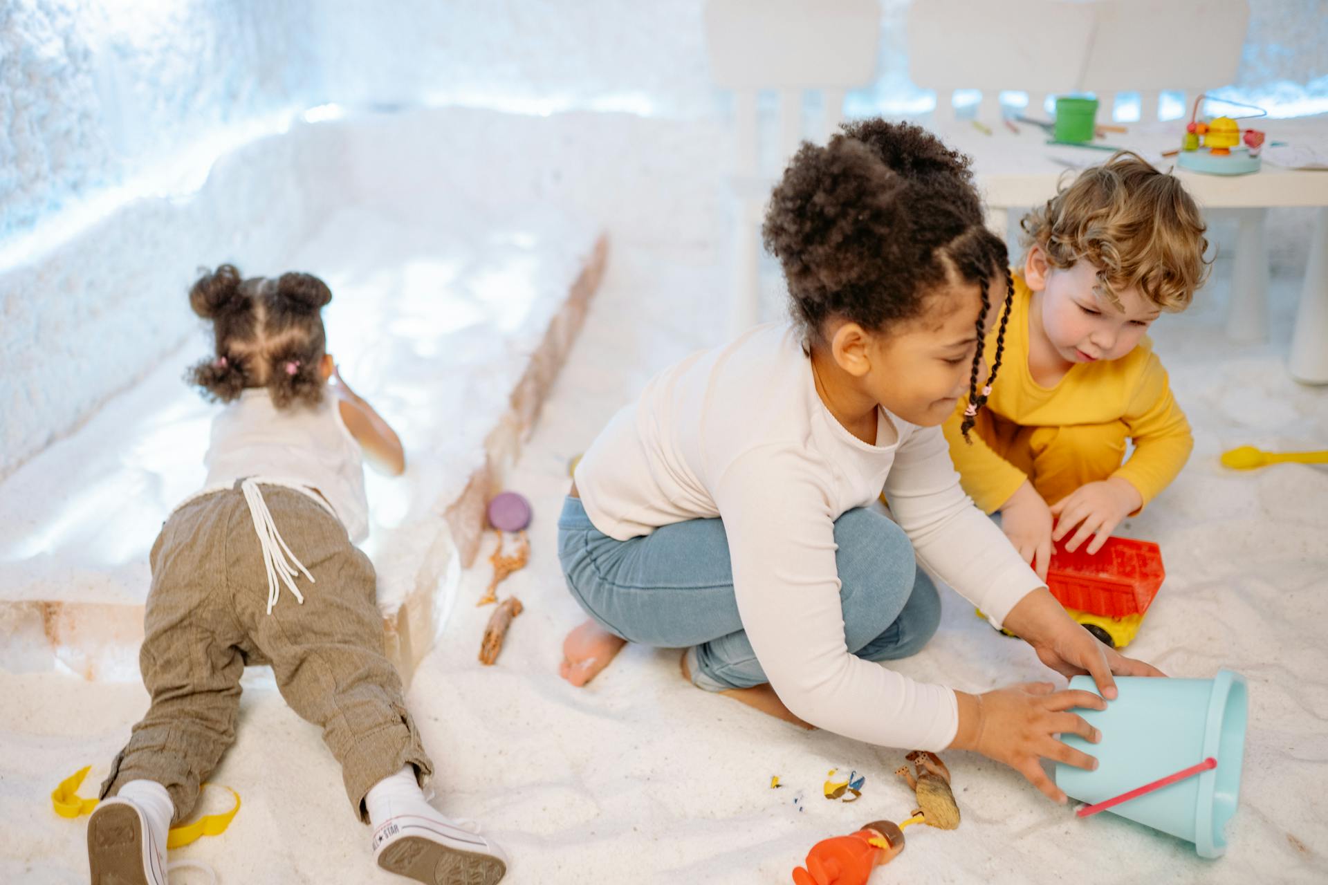 Three children engaging in indoor play with toys and a bucket, surrounded by white sand.