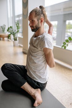 A bearded man with a man bun stretches in a yoga pose indoors, promoting wellness.