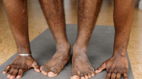 Close-up view of a man stretching on a yoga mat, performing a standing forward bend indoors.