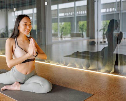 A young woman in yoga attire practices a seated yoga pose indoors by a reflective glass wall.