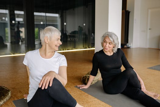 Two senior women smiling and stretching during a yoga class indoors, promoting fitness and togetherness.