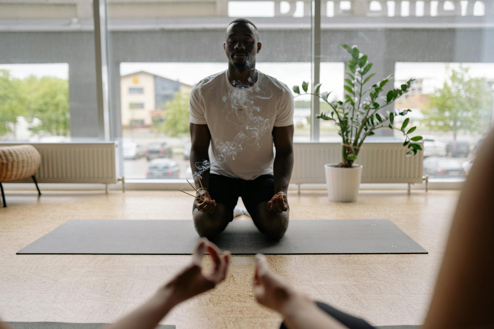 A man kneels in meditation indoors with incense smoke, surrounded by calm ambiance.