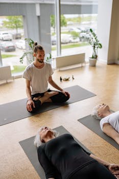 A calming yoga session with a teacher guiding two senior women in meditation on yoga mats in a sunlit studio.