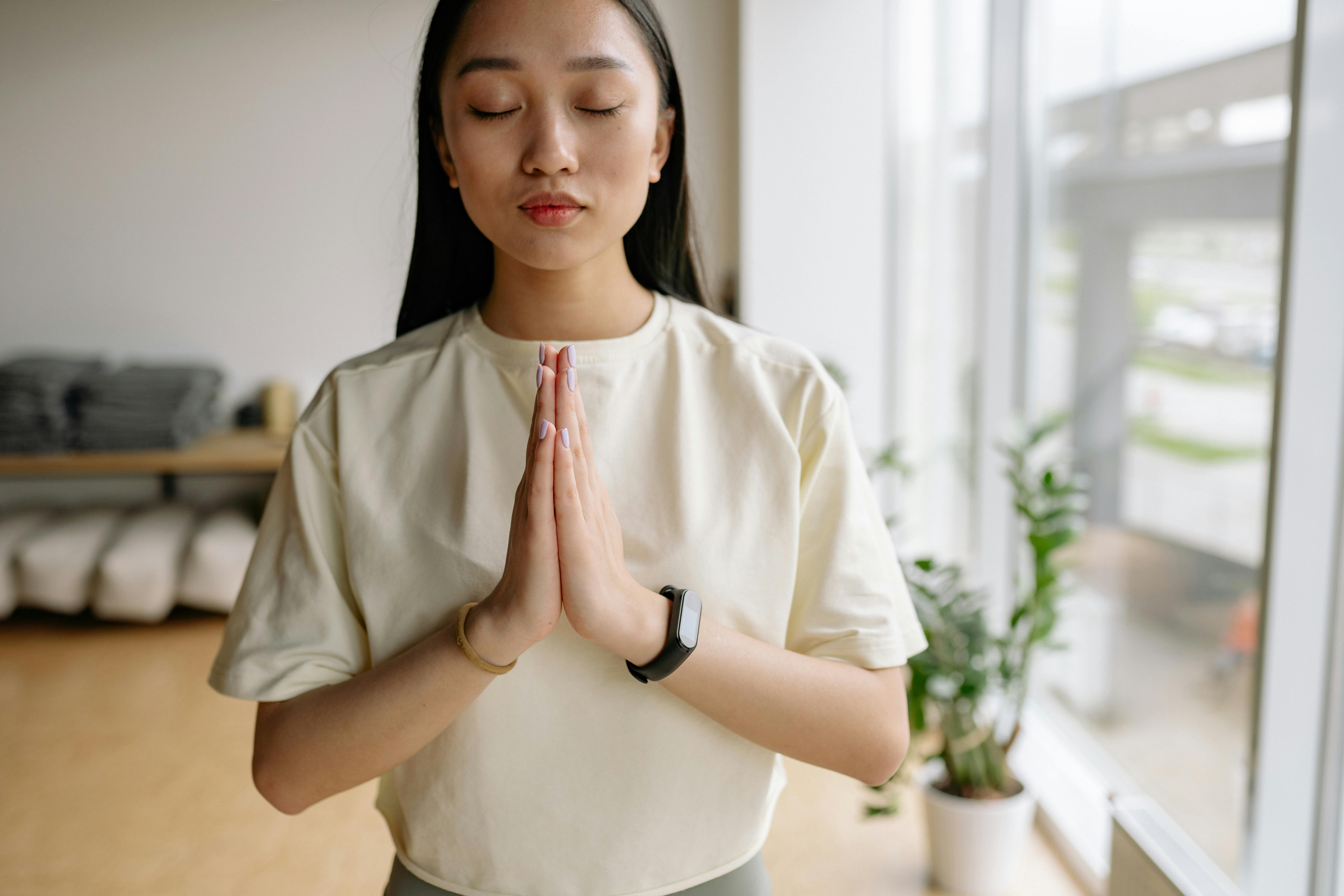 A person with closed eyes and hands pressed together in a prayer pose, standing in a studio with a window and a plant.