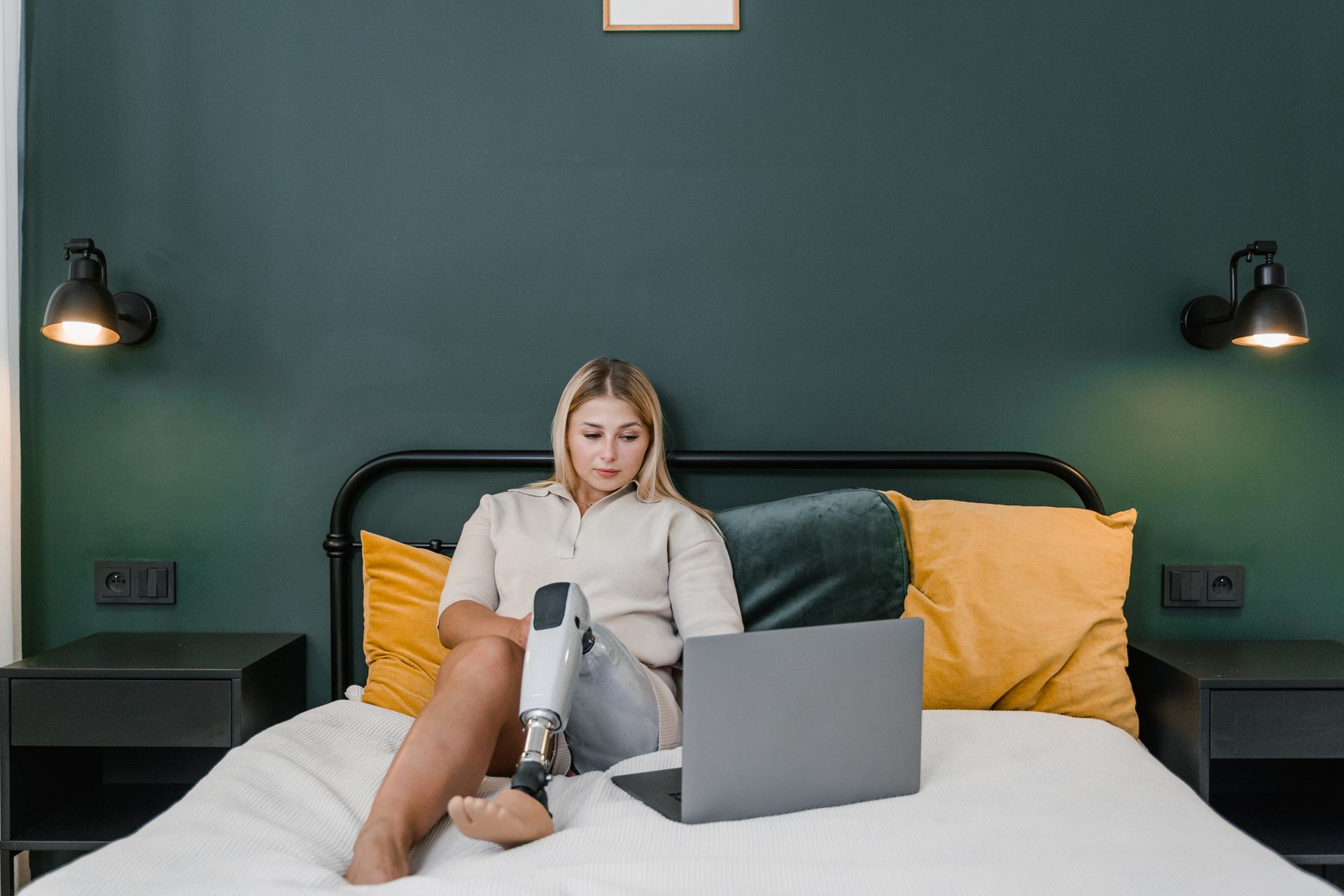 A woman with a prosthetic leg working on a laptop in a modern bedroom setting.