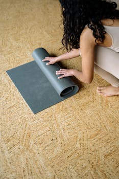 A woman rolls out a gray yoga mat on a wooden floor, preparing for a session.