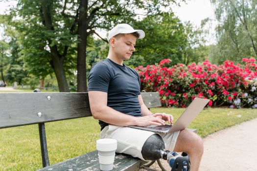 An adult male with a prosthetic leg working on a laptop while sitting on a bench in a park.