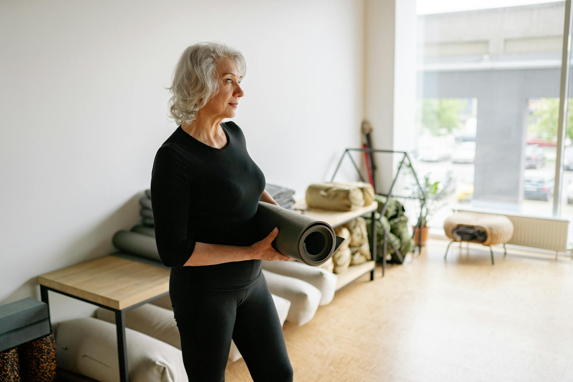 Elderly woman in black attire holding a yoga mat in a bright, minimalist space.