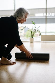 Elderly woman unrolling a yoga mat indoors, preparing for a calming workout.