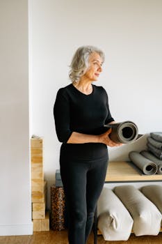 Elderly woman holding a yoga mat, standing in a bright indoor yoga studio.