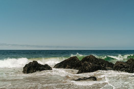 Peaceful view of ocean waves crashing on a rocky shoreline under clear blue sky.