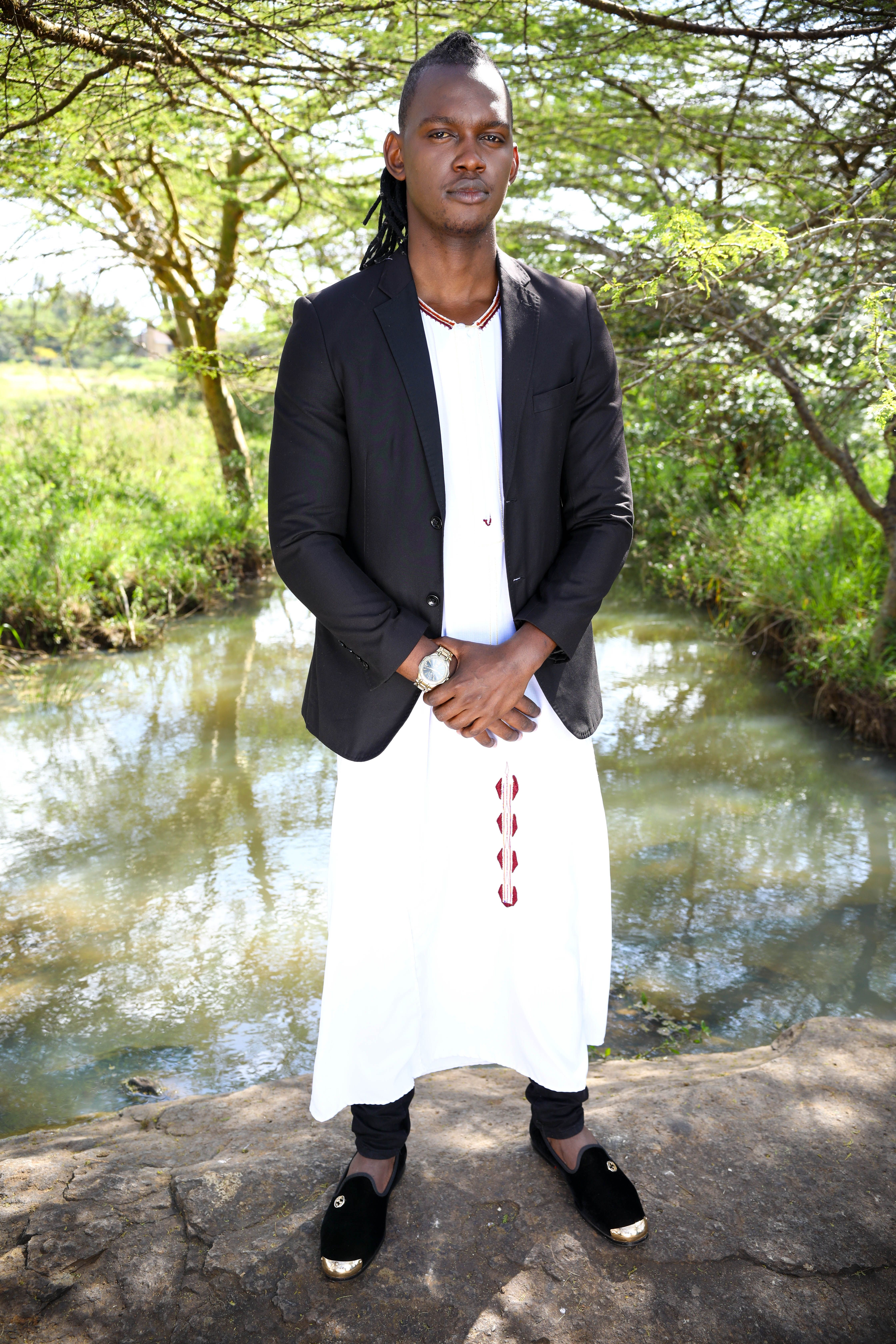 A man in a white traditional outfit and black jacket poses confidently by a stream.