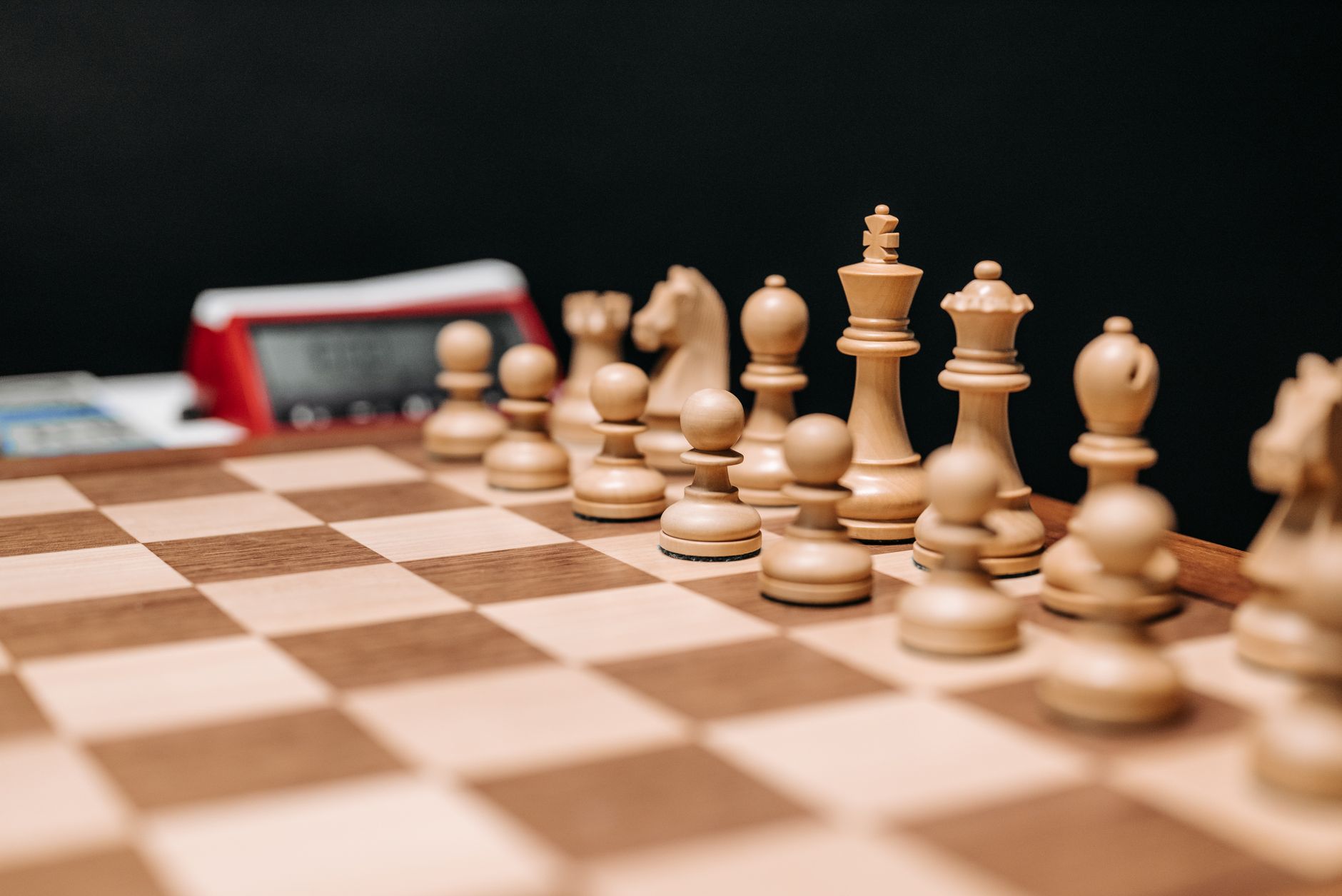 Detailed view of wooden chess pieces on a board with a chess clock in the background.