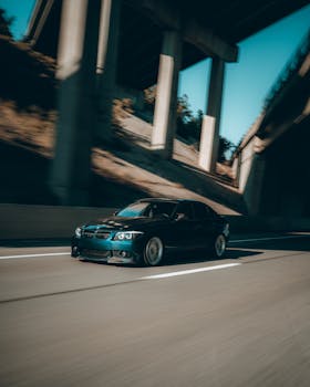High-speed black car driving on a sunny highway under an overpass, captured in a motion blur.