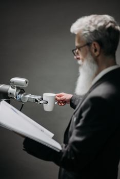 Elderly man interacts with robot arm holding a cup, showcasing innovation indoors.