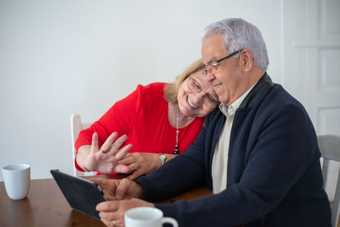 An older couple smiles and waves during a video call on a tablet at their kitchen table