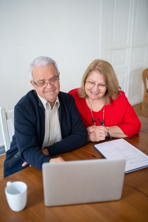 Free Senior couple sitting at a table, smiling while using a laptop indoors. Perfect for tech and lifestyle themes. Stock Photo