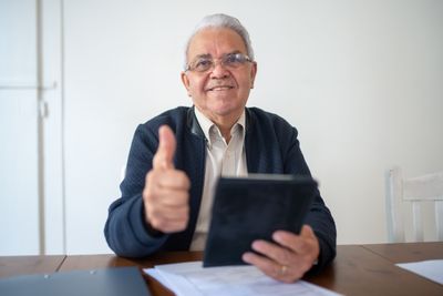 Smiling senior man seated with a tablet giving a thumbs up, expressing positivity.