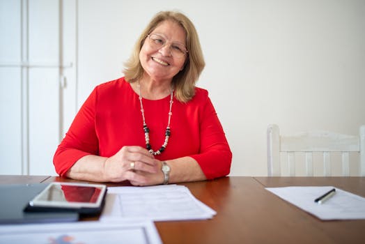 Warm and approachable senior woman sitting at a desk with documents, wearing a bright red top.