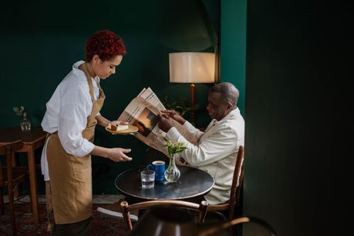 Free Waitress serving cheesecake to a man reading a newspaper in a cozy café setting. Stock Photo