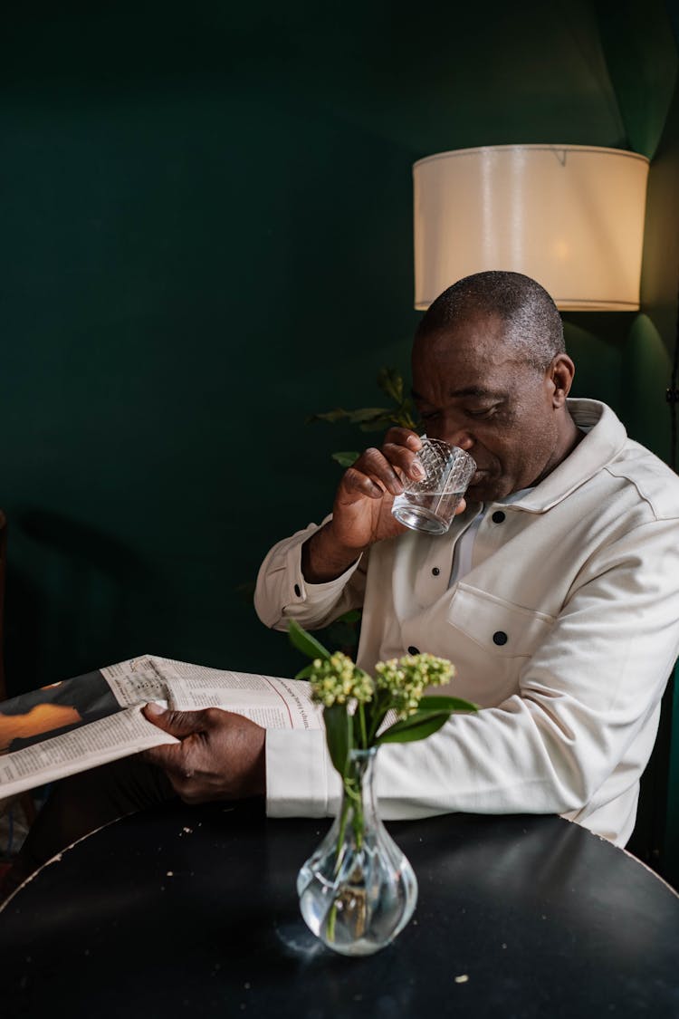 A Man Drinking A Glass Of Water While Reading The Newspaper