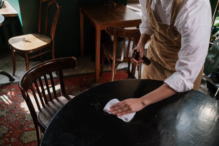 Person In White Shirt And Brown Apron Cleaning Black Table With White Wipe