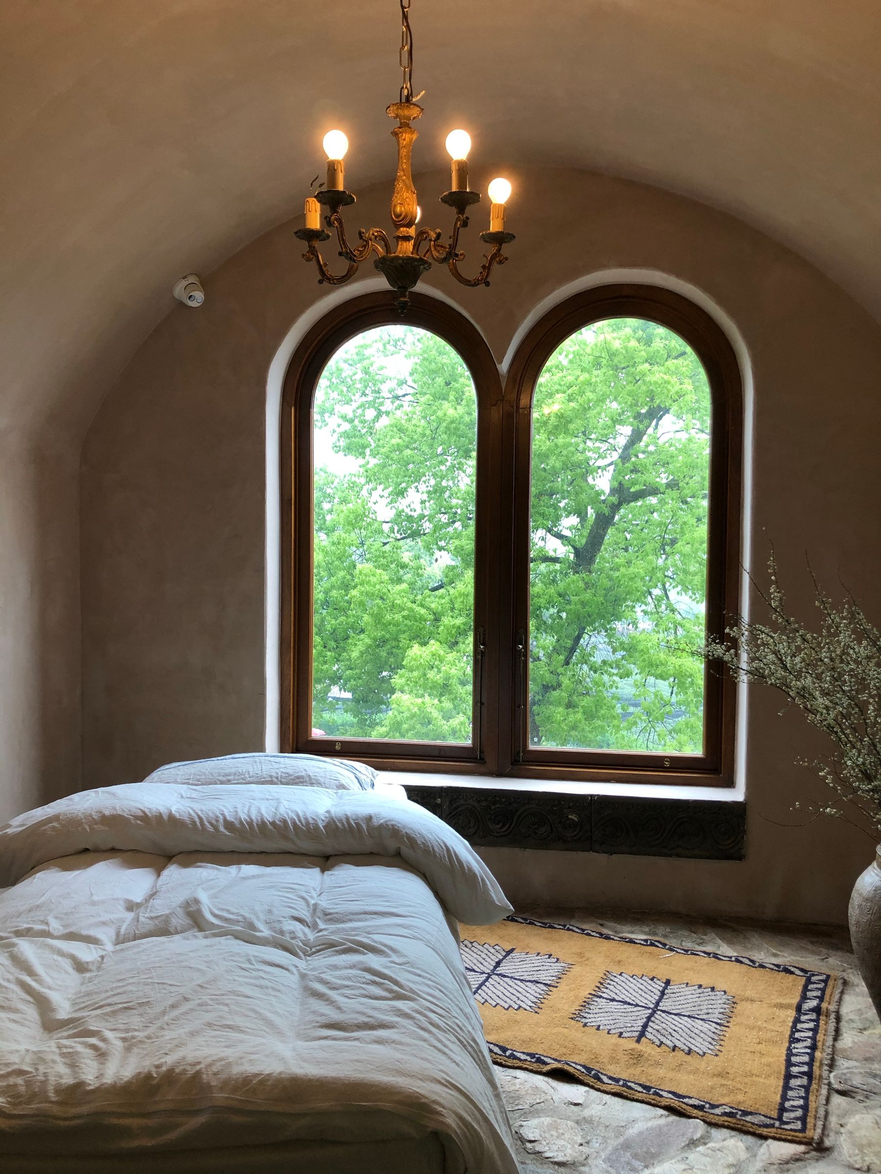 Tranquil bedroom with an arched wood-framed window, white bedding, woven rug, greenery and a rustic chandelier