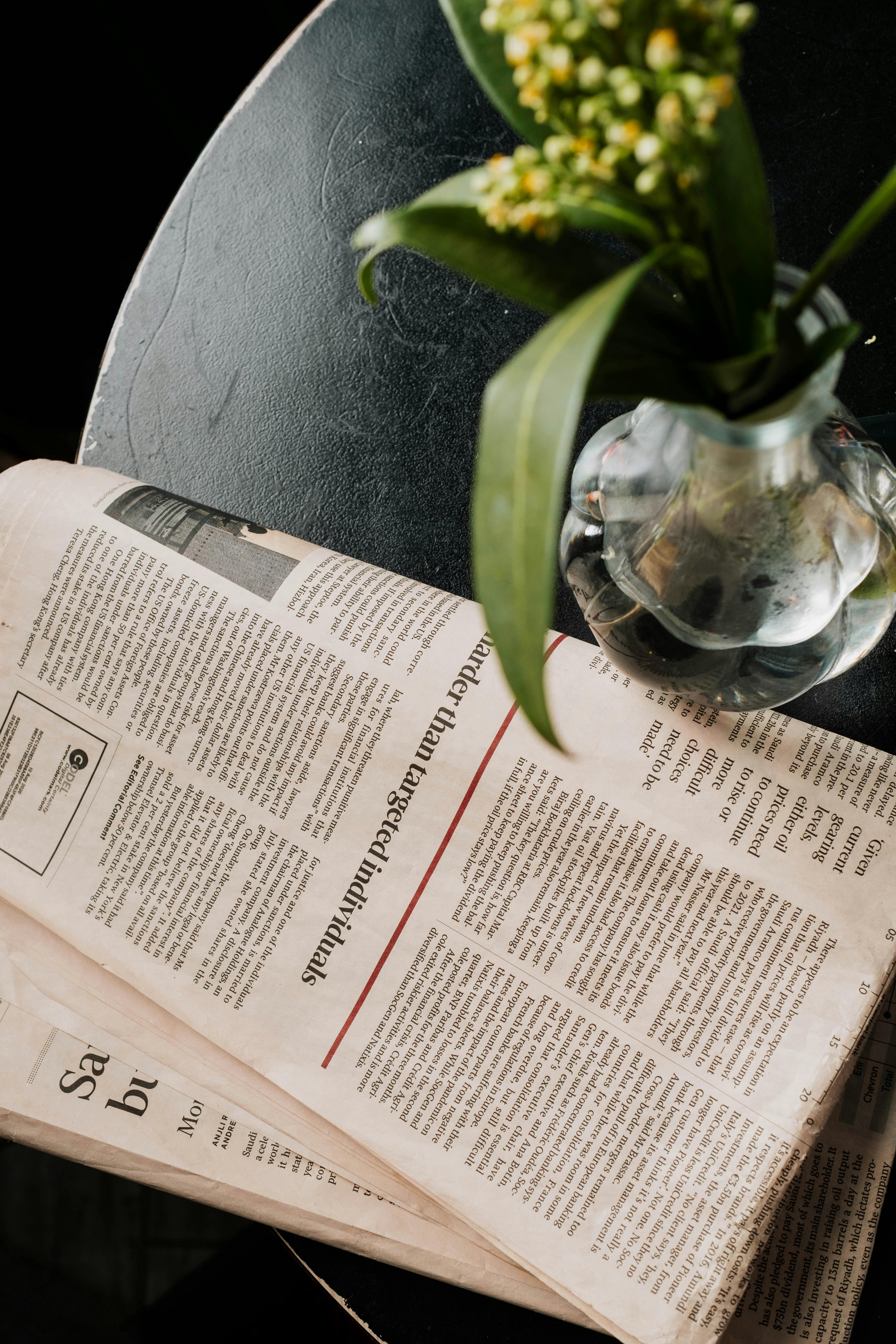 A top view of a glass vase with green leaves beside a folded newspaper on a dark table.