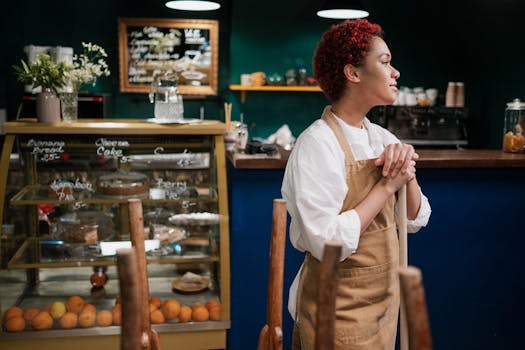 Waitress in a café wearing an apron stands thoughtfully by the counter with desserts on display.