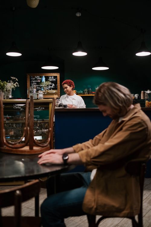 Free Barista with dyed red hair serves a customer in a dimly lit café, capturing a relaxed evening vibe. Stock Photo