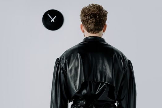 Back view of a man in a black leather jacket facing a wall clock indoors.