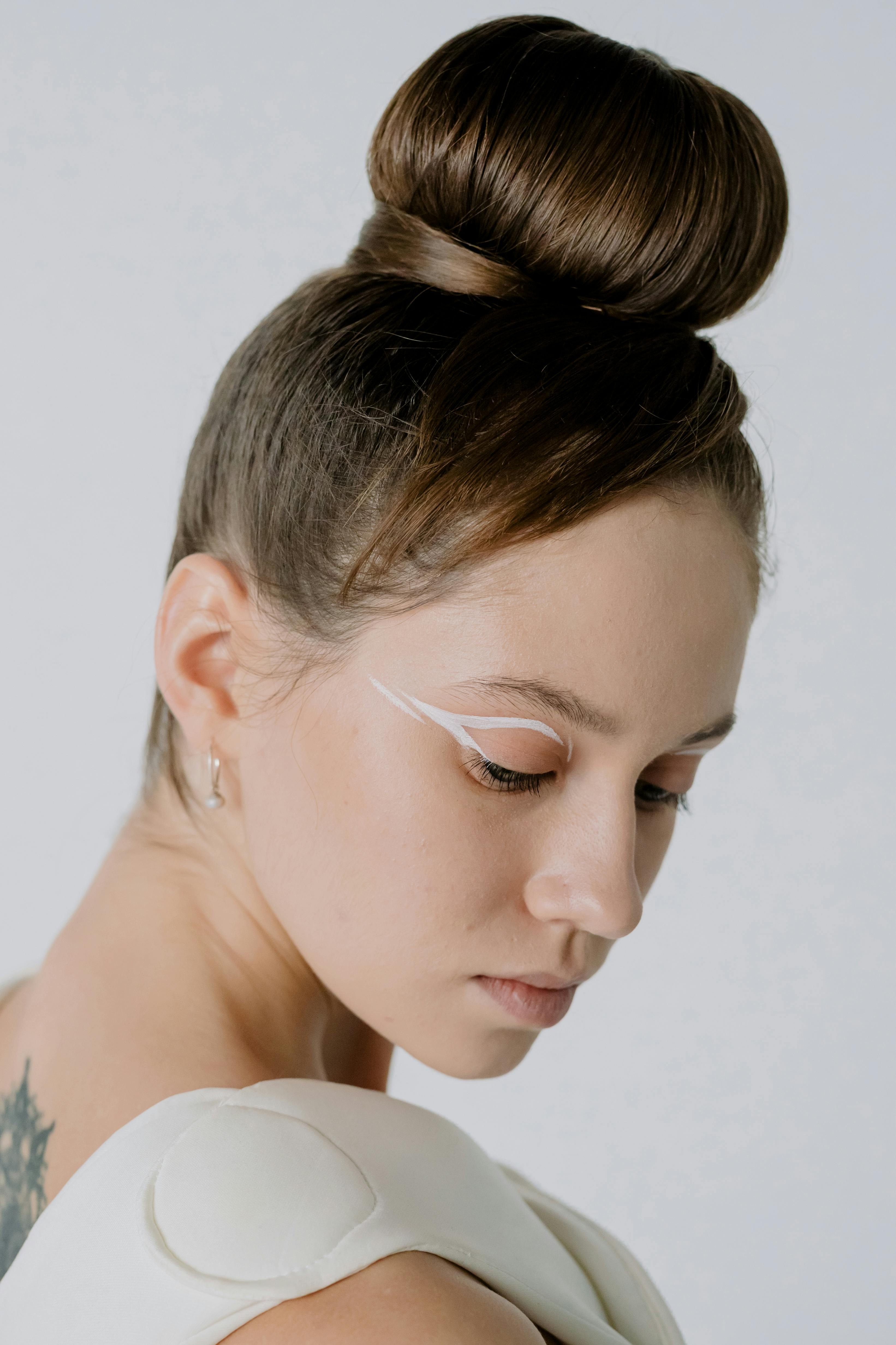Portrait of a woman with a fashionable hair bun, showcasing artistic makeup, captured in a minimal studio setting.