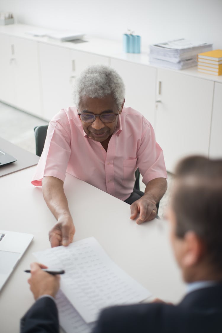 An Elderly Man In Pink Button-Up Shirt Sitting On A Chair While Handing A Paper To The Man Sitting In Front Of Him