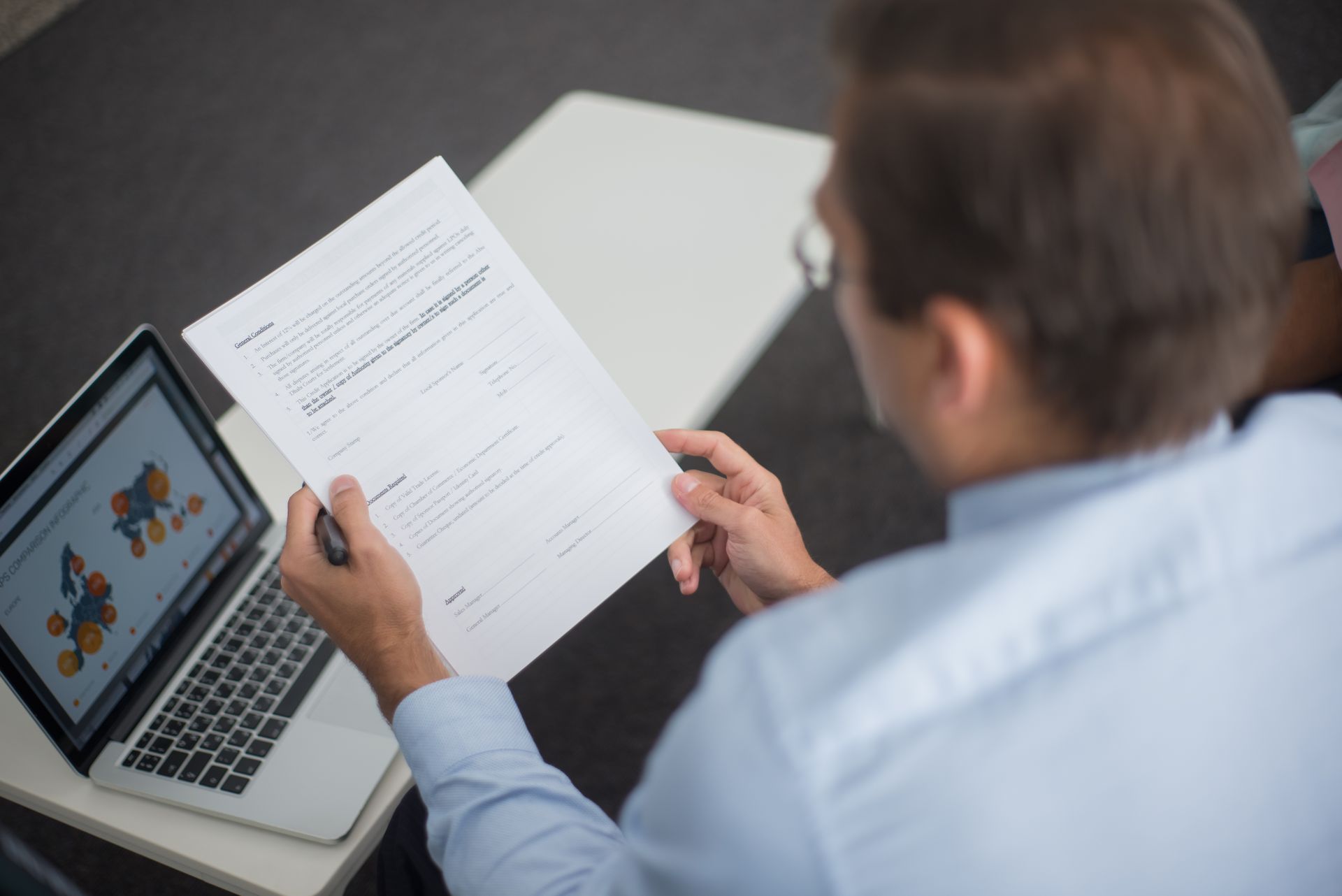 Professional in blue shirt reviewing documents next to laptop displaying charts.