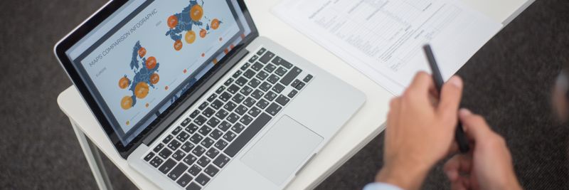 Professional analyzing data infographics on a laptop at an office desk, featuring documents and a pen.