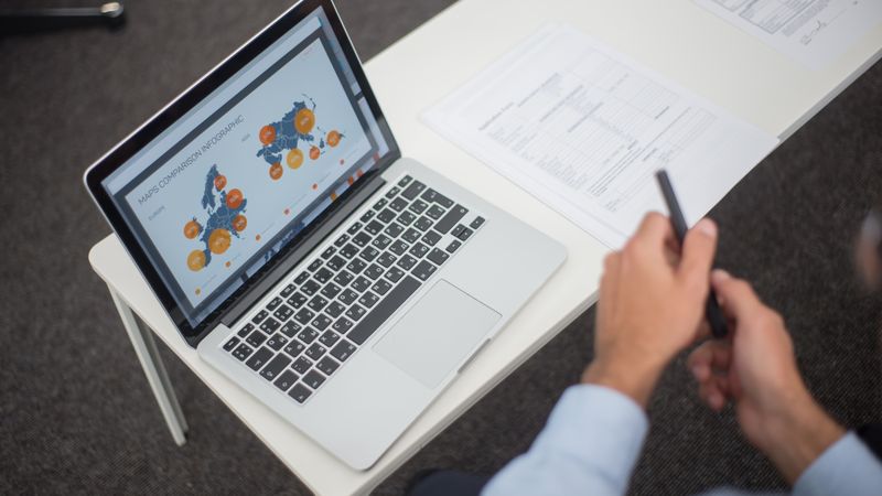 Professional analyzing data infographics on a laptop at an office desk, featuring documents and a pen.